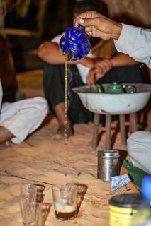 Person pouring tea from a blue teapot into glasses on sandy ground in a traditional desert