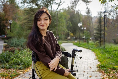 A woman with cerebral palsy with long dark hair sits comfortably on her walker in a sunlit park,