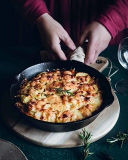 Hands arrange a sizzling dish of potato gratin topped with herbs in a black skillet. The wooden