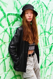 A young woman with long red hair and a black bucket hat poses against a vibrant green graffiti wall