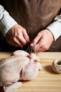A butcher skillfully ties a raw chicken with twine on a wooden table, demonstrating traditional