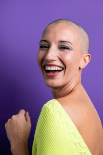 A vibrant portrait of a middle aged woman with a shaved head, smiling brightly against a purple