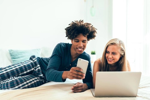 A smiling couple lounging on a bed while engaging with technology. The man shows something on his