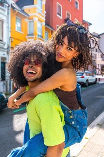 Young man giving piggyback ride to his girlfriend, both wearing sunglasses and smiling, enjoying a