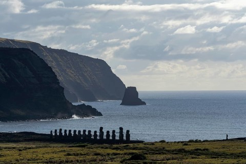 Moai statues of Ahu Tongariki in a row on the coast, behind steep cliffs and rock formation in the
