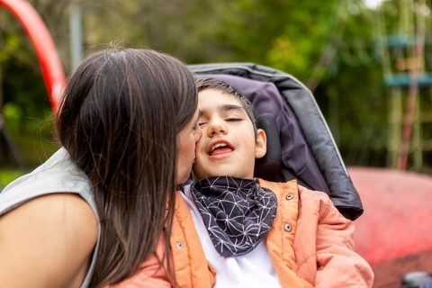 A tender moment as a mother kisses her son with cerebral palsy on the cheek. The son, seated in a
