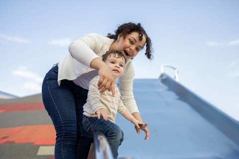 A joyful mother assists her baby on a playground slide in a sunny park. The image captures the