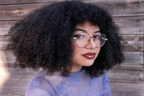 A confident young Hispanic Gen Z woman with curly hair and glasses gazes directly at the camera.