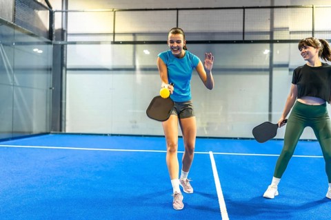 Two young women are playing pickleball on an indoor court, smiling and moving with paddles and a