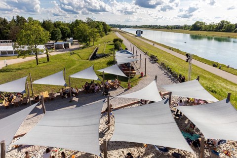 People relaxing under awnings by a river running through a green park, Oberschleißheim regatta