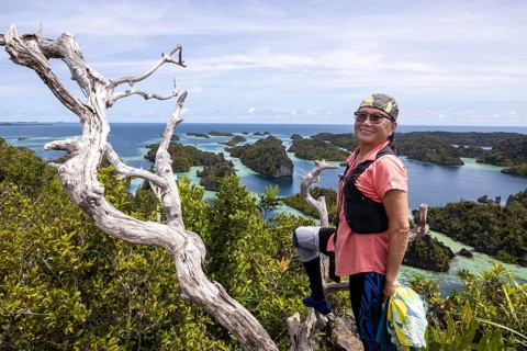 A person stands triumphantly on a cliff, enjoying breathtaking views of a lush Raja Ampat