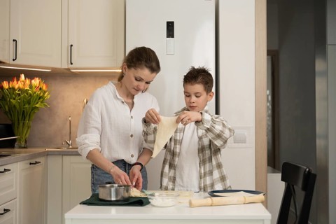 A mother helps her son with baking in a cozy kitchen. The boy is stretching dough, while the mother