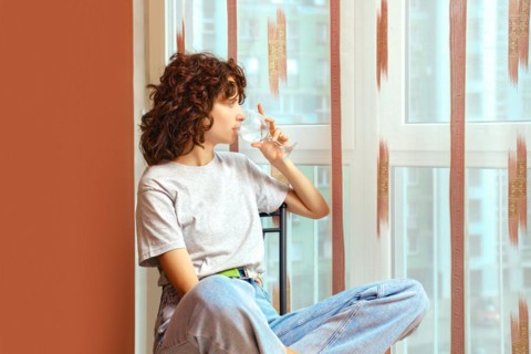 Woman sits by window, drinks mineral water and listens to sound of rain outside window