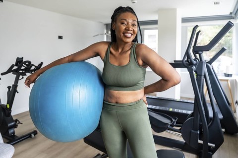 Happy woman wearing sportswear, leaning on a fitness ball in a hotel gym, promoting a healthy