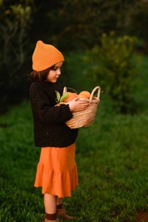 A young child in a knitted orange hat holds a basket full of oranges, standing on lush green grass
