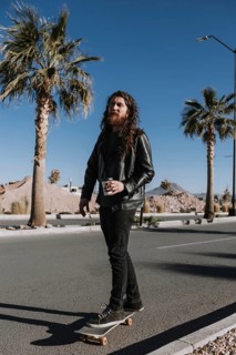 A man with long hair and a beard skateboards on a sunny street, holding a coffee cup. He wears a