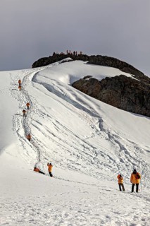 People in orange jackets climb a snow-covered mountain, tourists hike across the ice and rocky