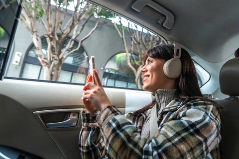 Woman wearing headphones and plaid jacket riding in the back seat of a car, engaging with her