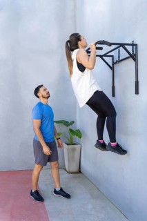 Determined woman performing pull-ups on a wall-mounted bar while personal trainer observes and
