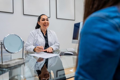 Aesthetic doctor in lab coat sitting at a desk, smiling and writing notes while consulting a