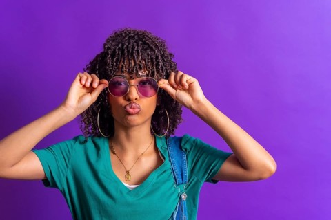 Fashionable young woman with curly hair adjusting her purple sunglasses and pouting on a vibrant