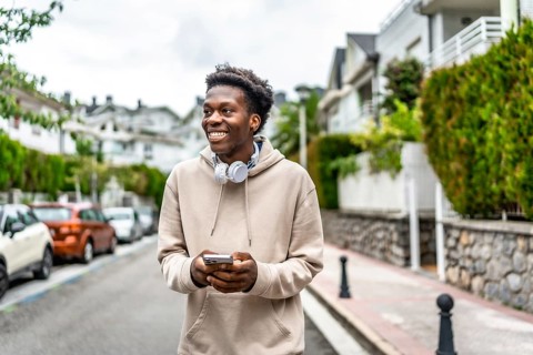 African cool and smiling man using mobile phone while walking along a residential street
