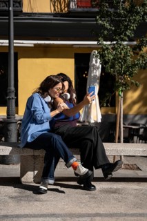 A multiethnic lesbian couple sits on a bench, sharing a joyful moment taking a selfie under the sun