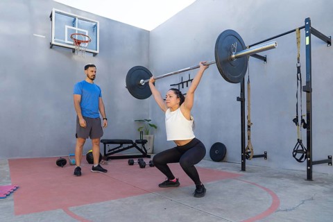 Young woman is performing squats with a barbell in an outdoor training area, under the supervision