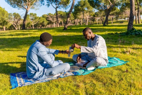 Two senegalese men in traditional dashiki clothing are sitting on a blanket in a park, pouring and