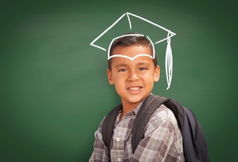 Young Hispanic Student Boy Wearing Backpack Front Of Blackboard with Graduation Cap Drawn In Chalk