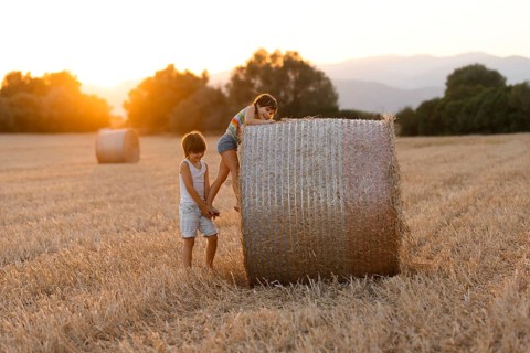 Two siblings, a boy and a girl, play near a large haystack in a field bathed in golden sunset