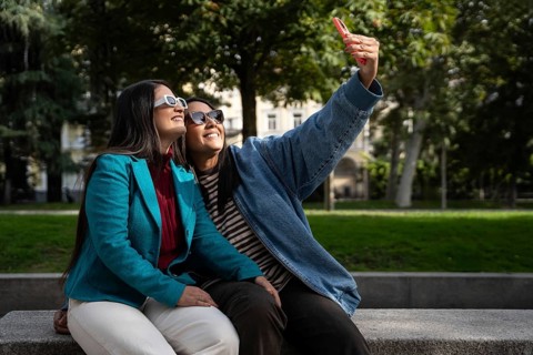 A joyful Latin lesbian couple in sunglasses takes a selfie in a sunny park. They are sitting on a