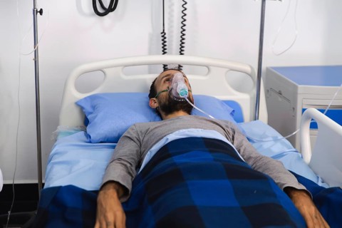 A patient lies in a hospital bed using an oxygen mask for respiratory support. Various medical