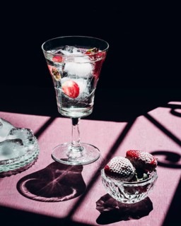 A refreshing gin tonic with ice cubes and strawberries sits elegantly on a pink table. Sunlight and