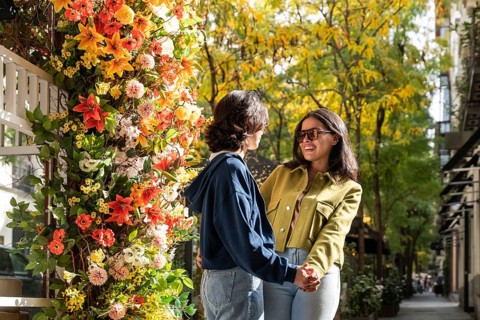 A lesbian couple stands hand in hand beside a wall of colorful flowers. They share a joyful moment