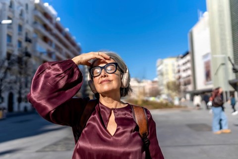 Mature woman with gray hair wearing headphones and eyeglasses, shielding her eyes from the bright