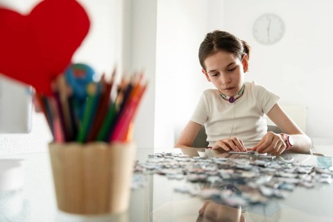 A young disabled girl with a tracheostomy focuses on assembling a puzzle at a table. Colored