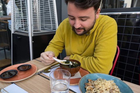 A man in a yellow sweater enjoys a traditional Asian meal at an outdoor restaurant table. He uses a