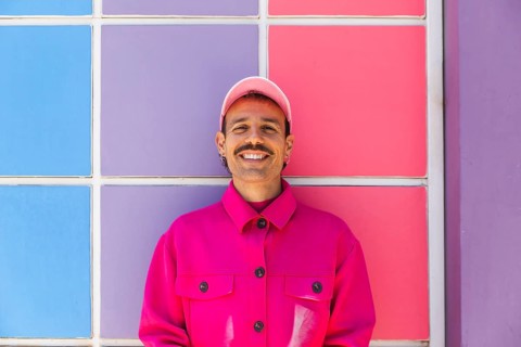 A young Spanish man with a mustache, wearing vibrant pink clothing, smiles joyfully against a