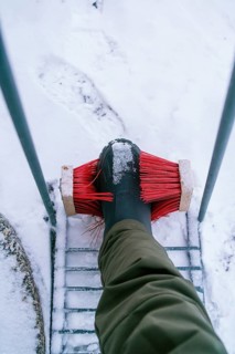 A person cleans snow off their boot using a red brush, set against a snowy backdrop in Swedish