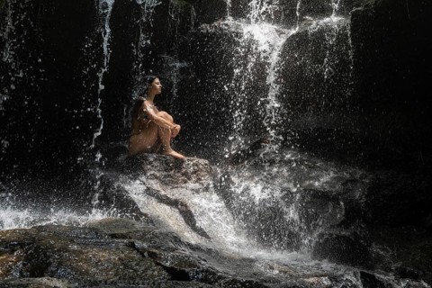 A serene scene of a woman sitting alone on rocks under a gentle waterfall, enveloped by nature of