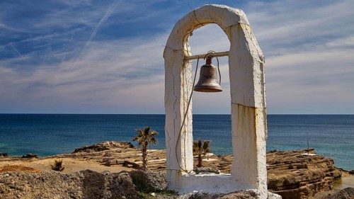A rusty bell hangs under a white arch by the sea in front of a clear sky and rocks, Chapel, Agios
