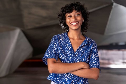 A cheerful woman with curly hair in a blue patterned shirt smiles confidently, standing with arms