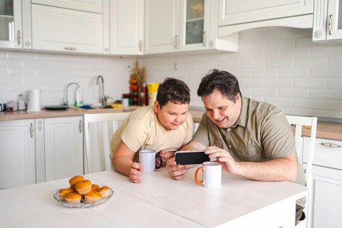 Father and son enjoy quality time in a cozy kitchen, sharing moments over a smartphone, with mugs