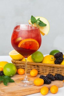 A glass of red sangria filled with citrus slices and mint sits on a wooden board. Surrounding the