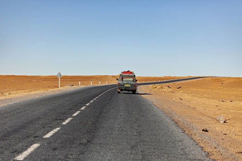 Lonely road with a vehicle in the distance, surrounded by empty sandy areas, under a clear sky, By