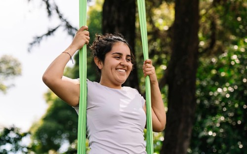 A latin woman joyfully practices aerial acrobatics on fabric in a lush Bogota park, surrounded by