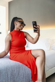 Smiling businesswoman wearing red dress using smartphone for video call while sitting on bed in