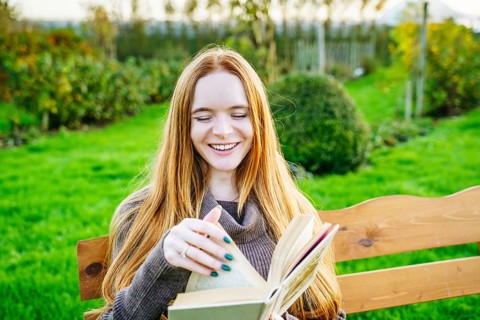 A beautiful pregnant woman with red hair enjoys reading a book on a wooden bench in a lush green