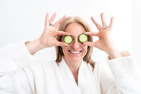 Mid-adult woman in a spa setting holding cucumber slices over her eyes, wearing a white robe. The